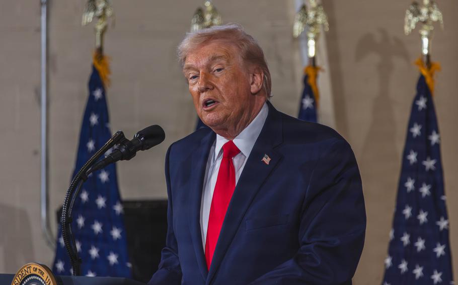 President Donald Trump in a dark suit and red tie speaks at a microphone in front of three U.S. flags.